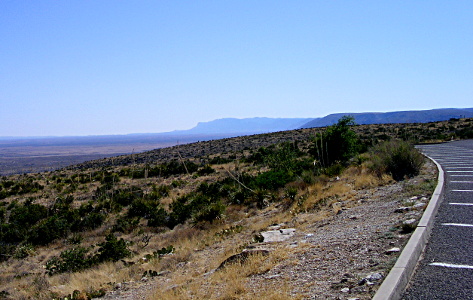 [The edge of the paved parking lot is on the right. The ground slopes away from the parking lot down a hillside with quite a bit of dark green shrubbery. In the far distance is a blue-grey outline of the top of a mountain range which then slopes sharply down to the horizon line. The sky is blue without any clouds.]